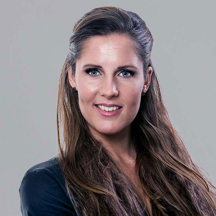 A portrait of a person with long, wavy hair, wearing a black jacket against a neutral background. Portrait of Marjanne Vroom.