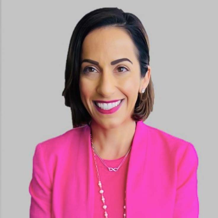 A person is wearing a pink blazer and a necklace. The background is grey. Portrait of Patricia Kakridas.