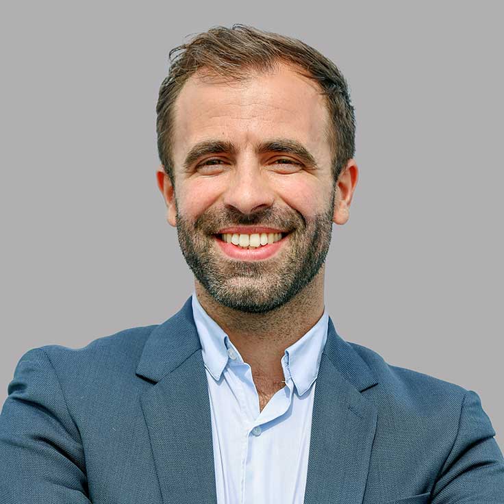 A man is wearing a blazer and shirt. He stands with crossed arms against a grey background. Portrait of Pierre-Emmanuel Burg.