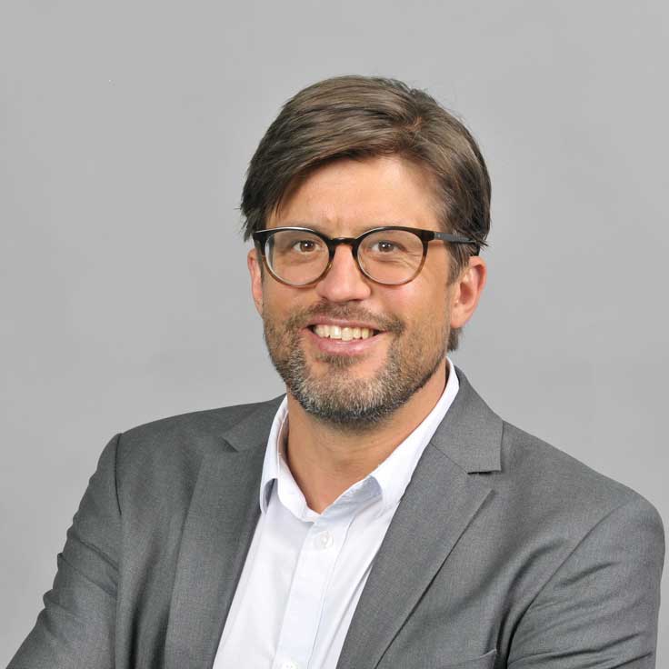 A man in a grey suit with a white shirt, looking towards the camera with his arms crossed. Portrait of Simon Lorenz.