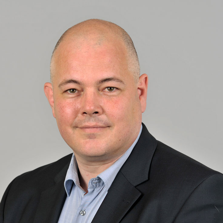 Ein Mann mit glänzender Glatze trägt einen schwarzen Anzug und ein hellblaues Hemd vor einem grauen Hintergrund. Portrait von Rob Hageman.