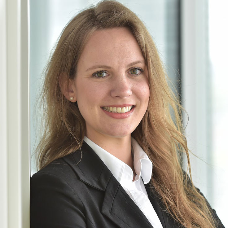 A person with long, wavy hair wearing a black suit and a white shirt leans against a wall. Portrait of Stephanie Buchloh.