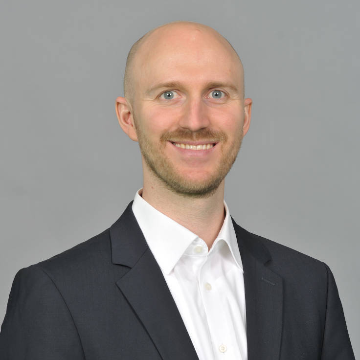 A man in a black suit and white shirt poses in front of a gray background, showing a professional appearance. Portrait of Alexander Möllmann.