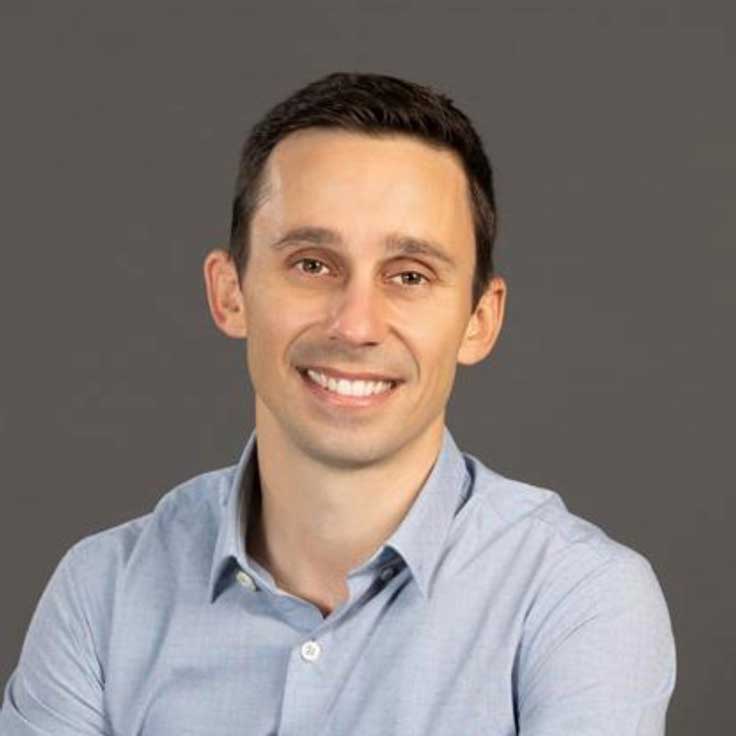 Ein Porträt eines Mannes mit einem hellblauen Hemd. Der Hintergrund ist grau, und er hat eine entspannte Pose eingenommen. Portrait von Andrea Baldi.