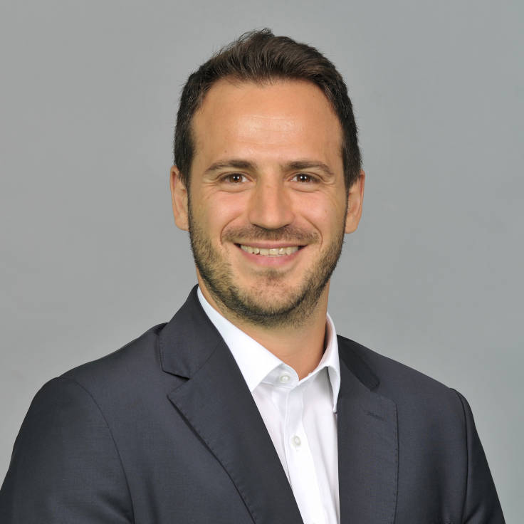 Ein Mann in einem dunklen Anzug und einem weißen Hemd posiert vor einem grauen Hintergrund. Portrait von Cihan Sönmez.