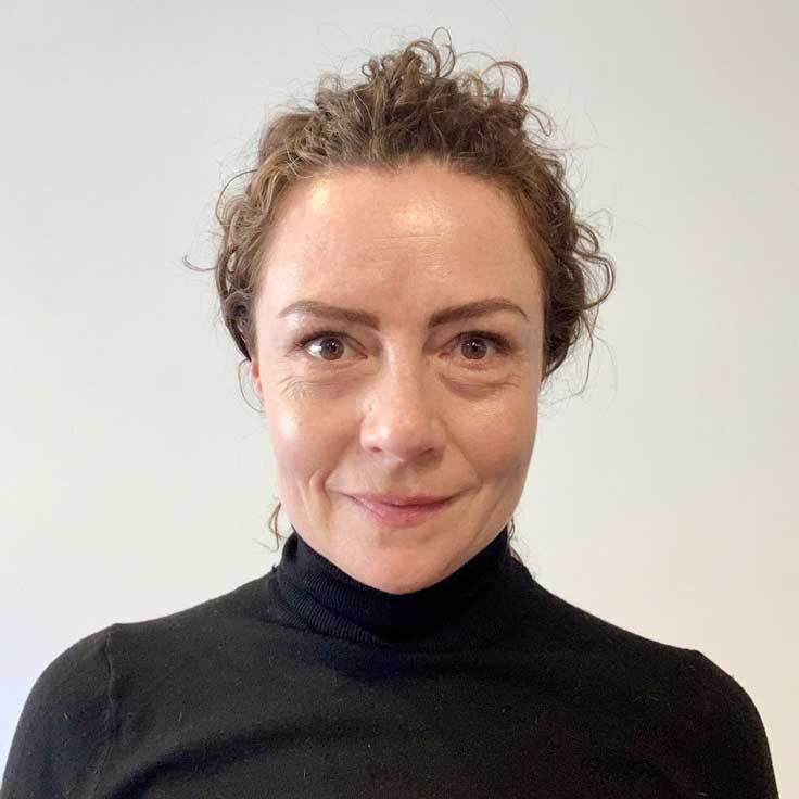 Eine Person mit lockigem Haar trägt einen schwarzen Pullover. Der Hintergrund ist hell und neutral. Portrait von Erika Vasquez.