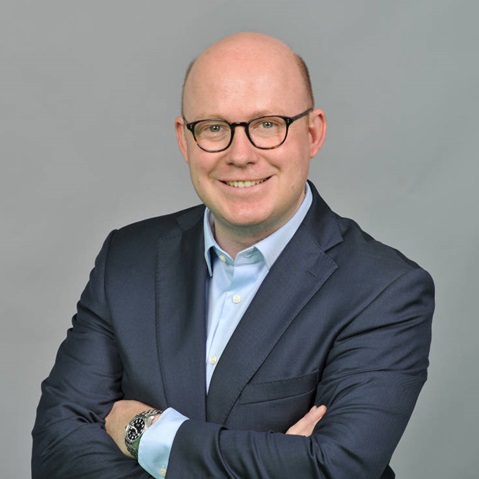 Ein Mann in einem dunklen Anzug mit gekreuzten Armen steht vor einem grauen Hintergrund. Portrait von Felix Christoph Ringenberg.