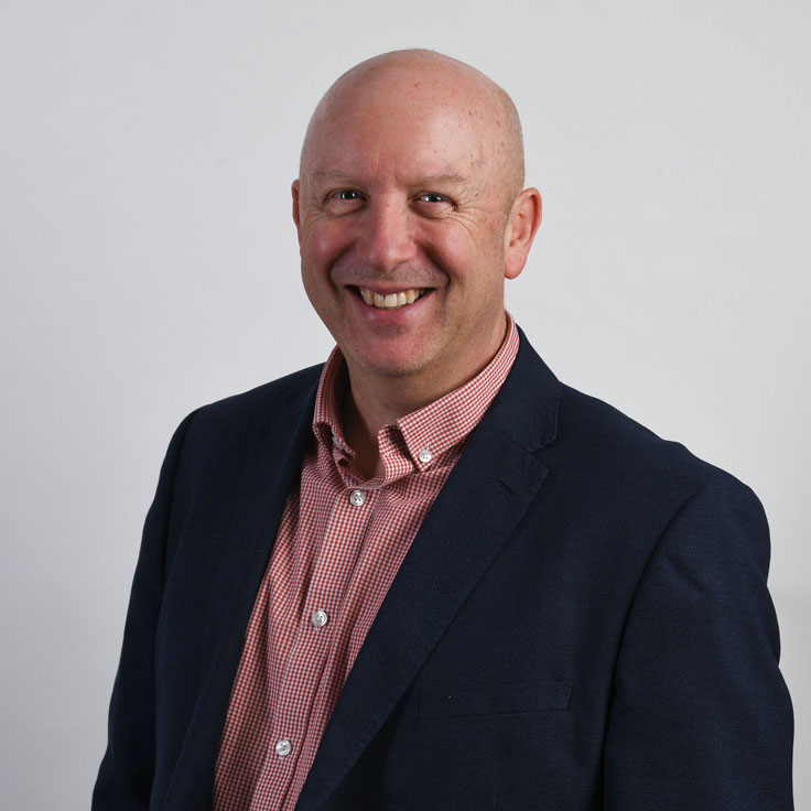 Ein Mann trägt einen marineblauen Blazer über einem karrierten Hemd und posiert vor einem schlichten Hintergrund. Portrait von Jeremy Smith.