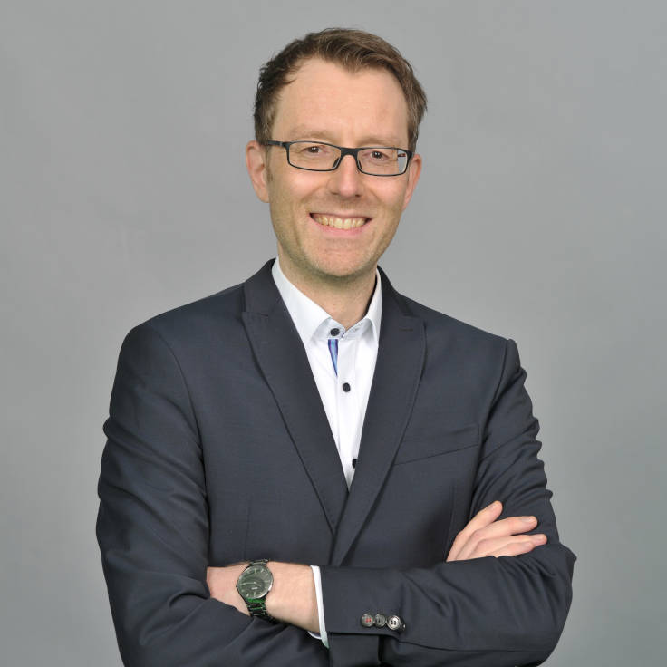 Ein Mann in einem Anzug steht mit verschränkten Armen vor einem grauen Hintergrund und strahlt Professionalität aus. Portrait von Kai Stevens.