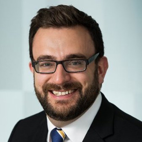 Eine Person mit kurzem dunklem Haar in schwarzem Sakko, weißem Hemd und blau-gelb gestreifter Krawatte vor hellem grauem Hintergrund. Portrait von Nick Stapley.