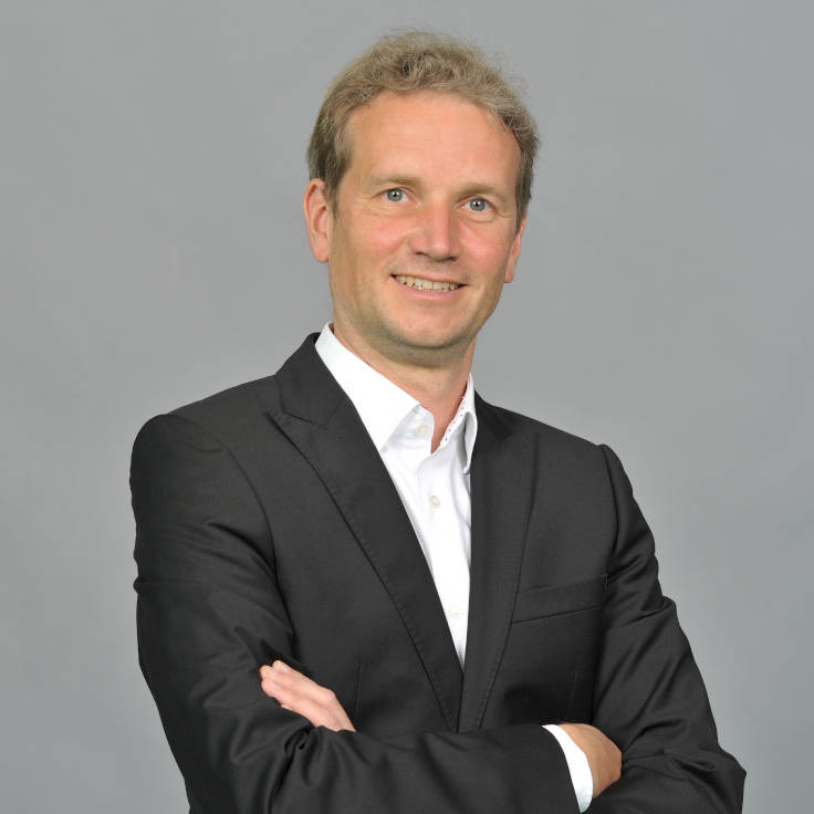 Eine Person in einem schwarzen Anzug mit verschränkten Armen, die vor einem grauen Hintergrund steht. Portrait von Nils Weise.