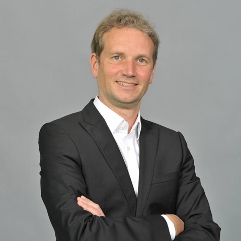 Eine Person in einem schwarzen Anzug mit verschränkten Armen, die vor einem grauen Hintergrund steht. Portrait von Nils Weise.