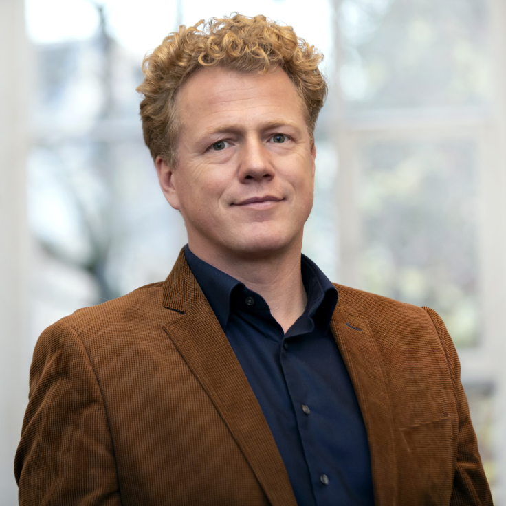 A man with curly blonde hair in a brown cord jacket and a navy shirt stands in front of large windows.