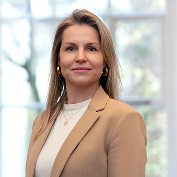 Eine Frau mit langen Haaren trägt einen beigen Blazer und goldene Creolen. Sie steht vor einem hellen Fenster. Portrait von Sophie Witsenburg.