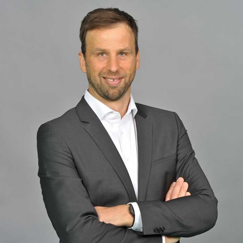 Ein Mann in einem grauen Anzug steht mit verschränkten Armen vor einem grauen Hintergrund. Portrait von Stefan Nass.