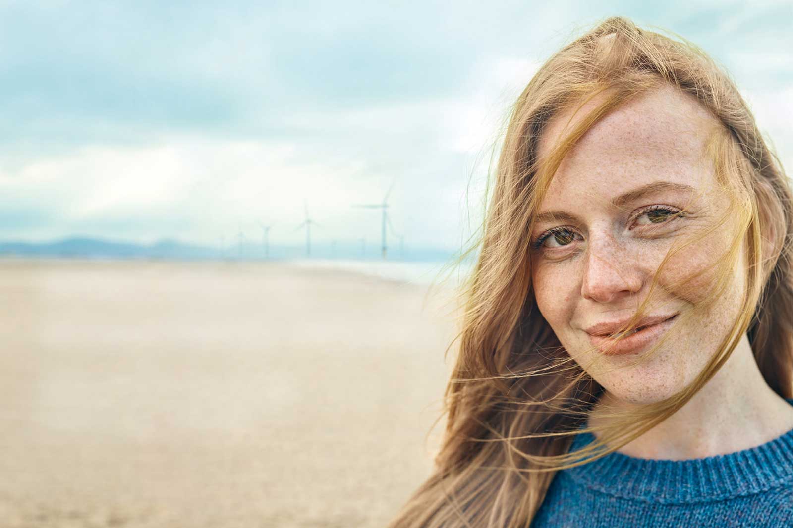 Eine Person mit langen, blonden Haaren vor einer Küstenlandschaft mit Windkraftanlagen im Hintergrund.
