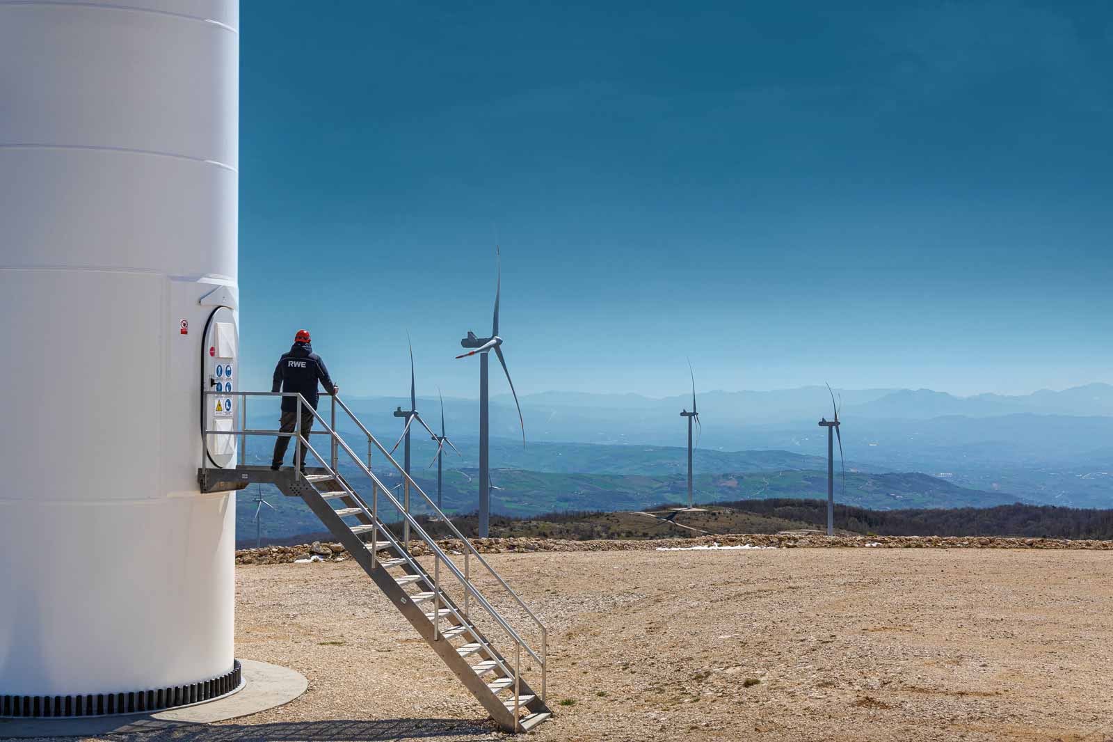 A technician stands on a ladder by a wind turbine. The background features several wind turbines and a mountainous landscape.