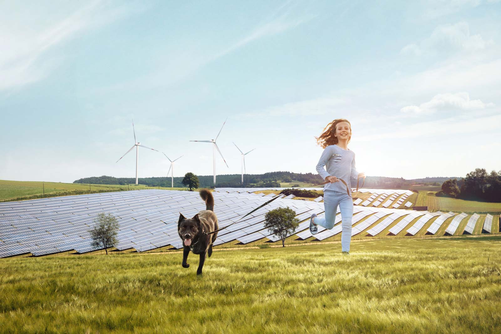 Ein Mädchen läuft fröhlich mit einem Hund in einem Feld neben Solarpanelen und Windkraftanlagen unter einem klaren Himmel.