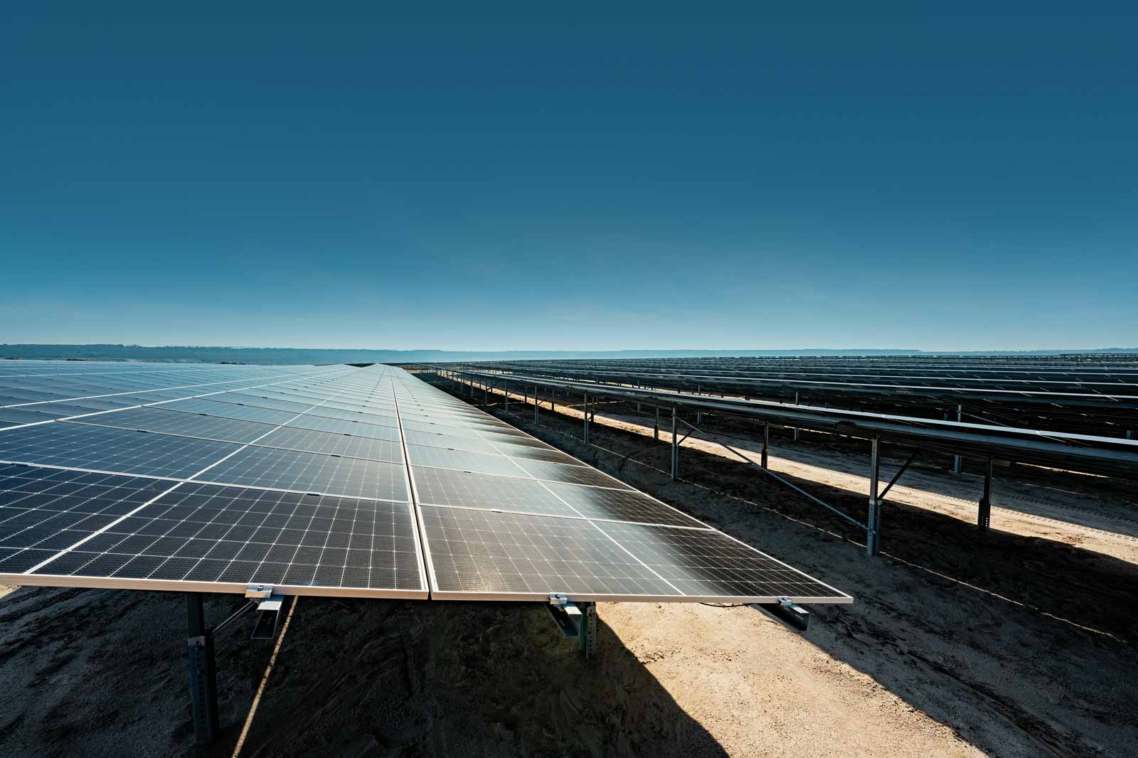 Ein Feld mit Solarpanels erstreckt sich unter einem klaren blauen Himmel. Der Boden ist trocken und sandig.