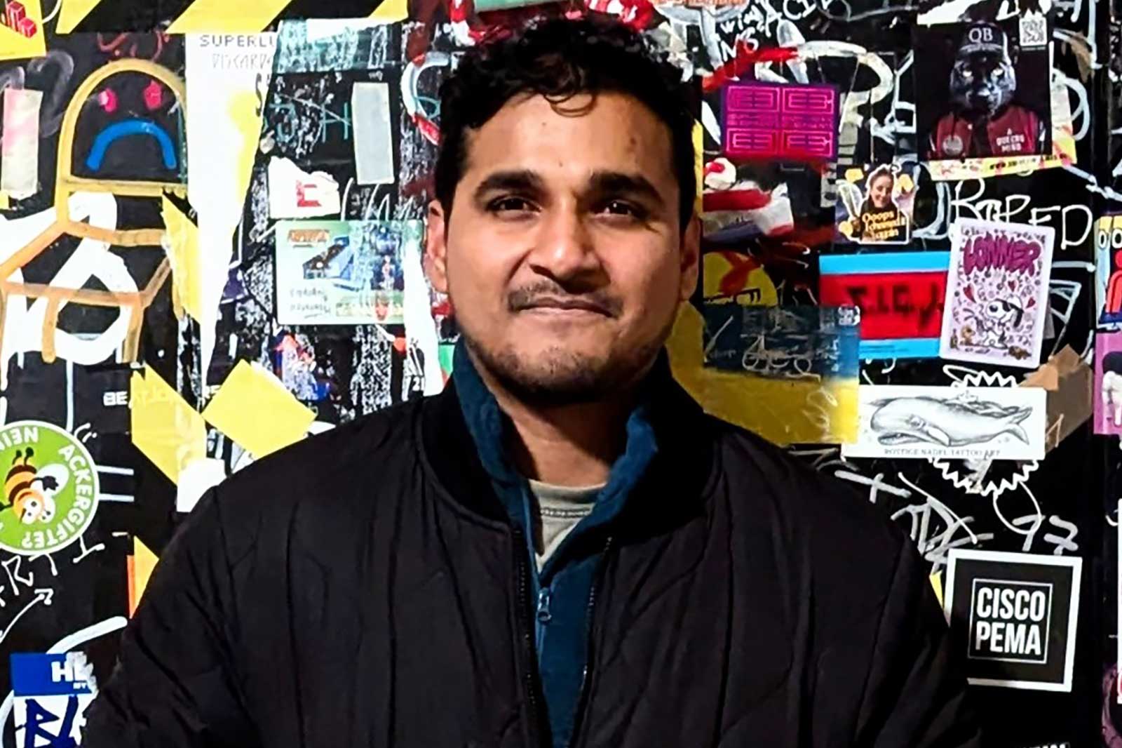 A man in a dark jacket stands against a colourful wall covered in stickers and graffiti.