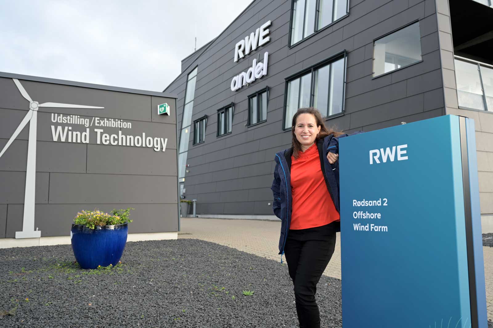 A person stands in front of a modern building featuring a sign about wind technology and a blue pot with plants.