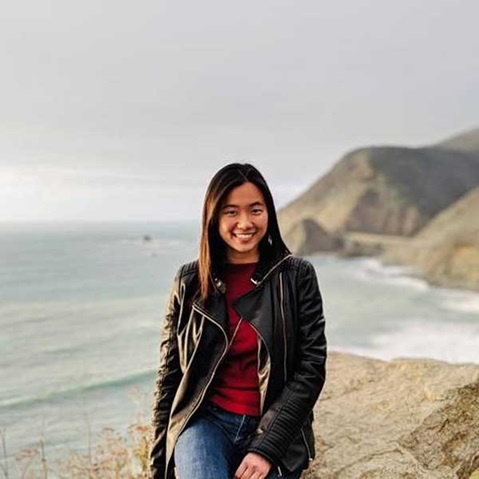 Eine Person sitzt am Ufer, trägt eine schwarze Lederjacke und blickt auf das Meer, während die Berge im Hintergrund sichtbar sind.