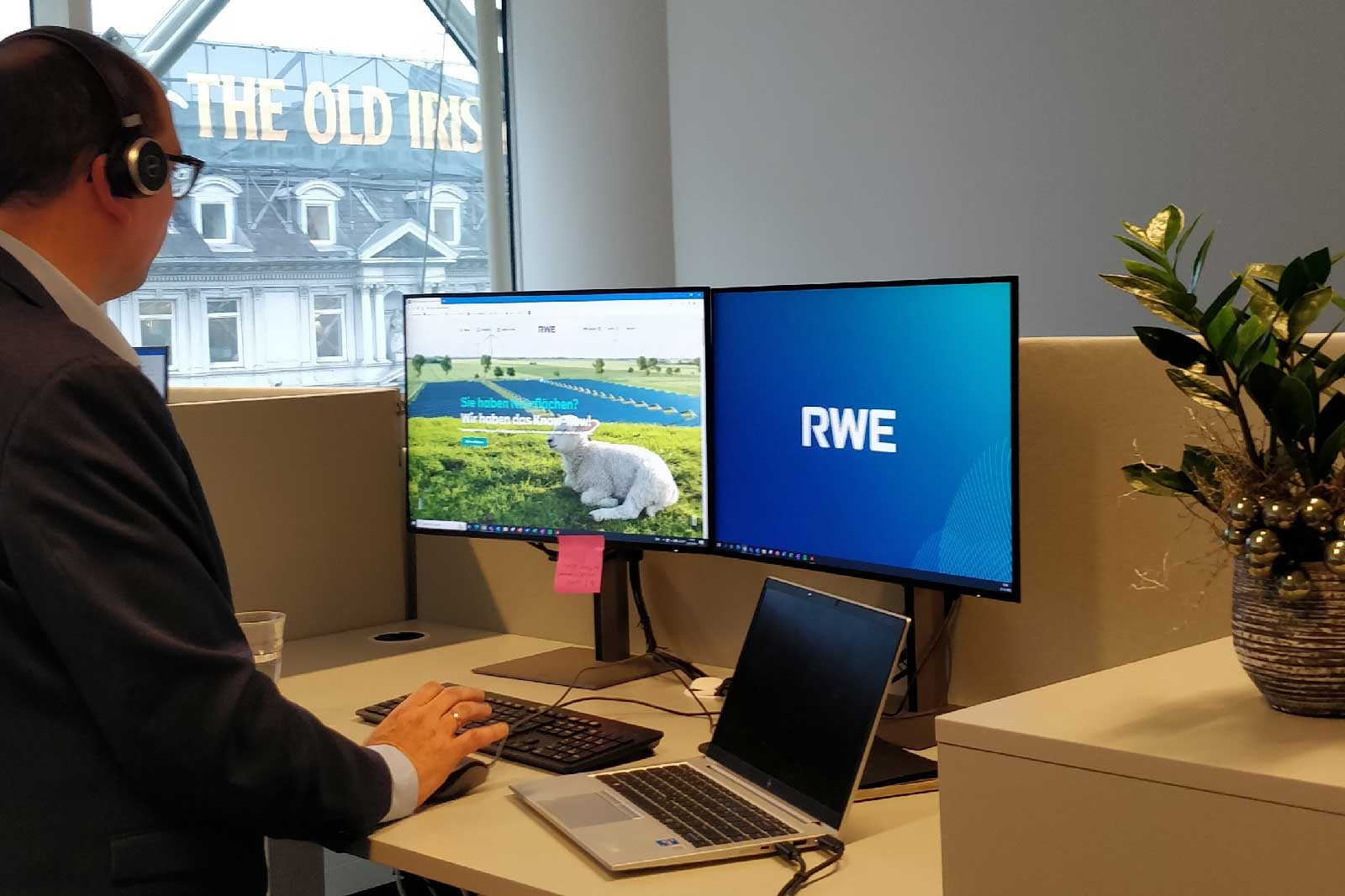 Ein Mann mit Kopfhörern arbeitet an zwei Bildschirmen in einem Büro mit einem Pflanzenarrangement.