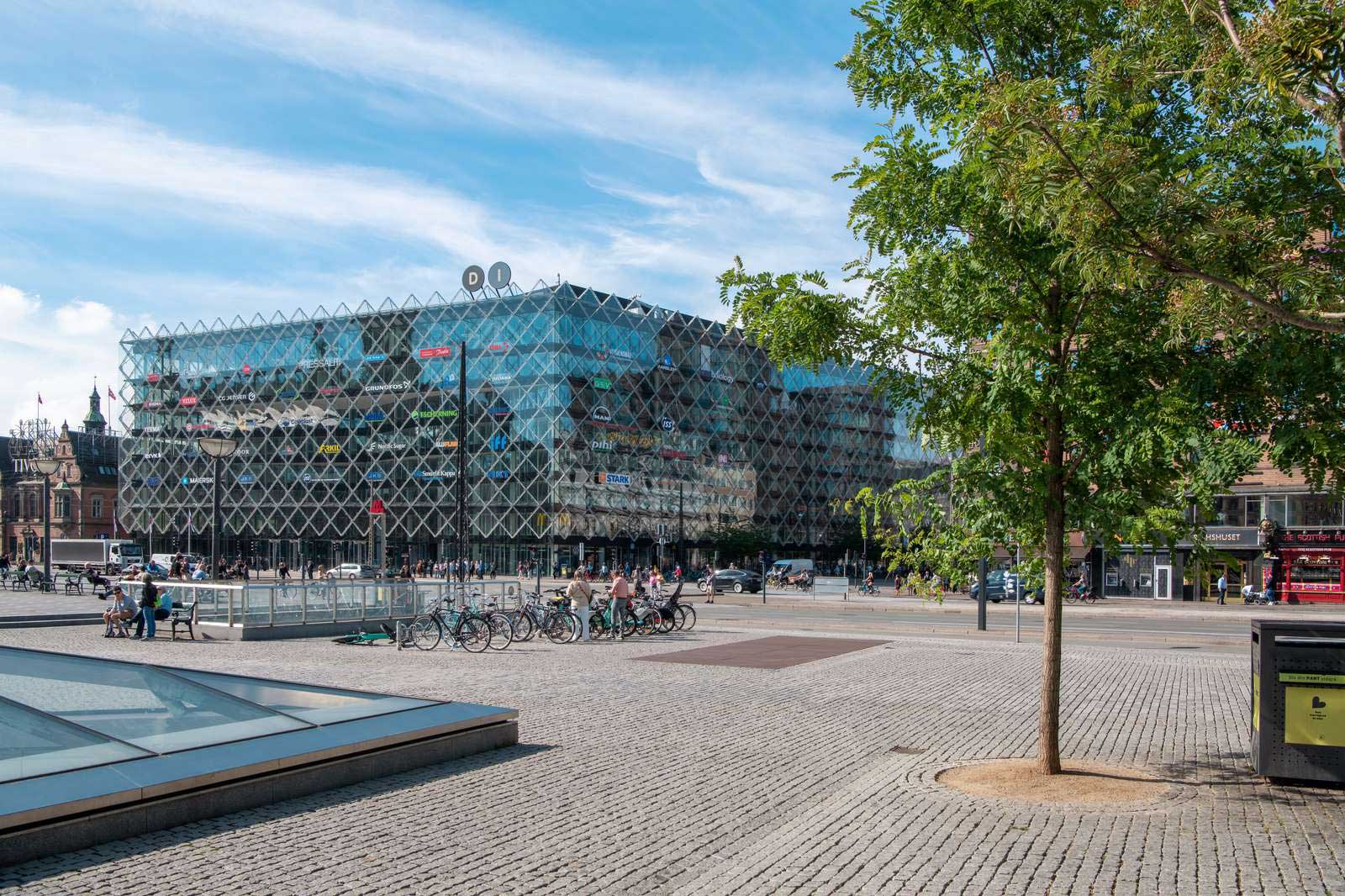 Moderne Glasgebäude mit einem auffälligen, dreidimensionalen Muster und einem Platz voller Fahrräder und Passanten.