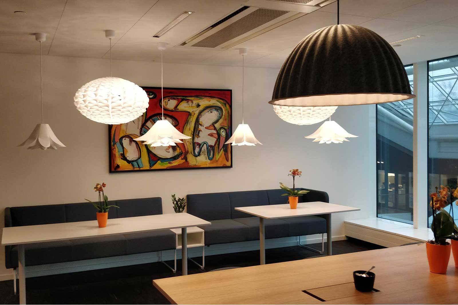 Ein modernes Besprechungszimmer mit stilvollen Pendelleuchten, einem bunten Gemälde und Topfpflanzen auf weißen Tischen.