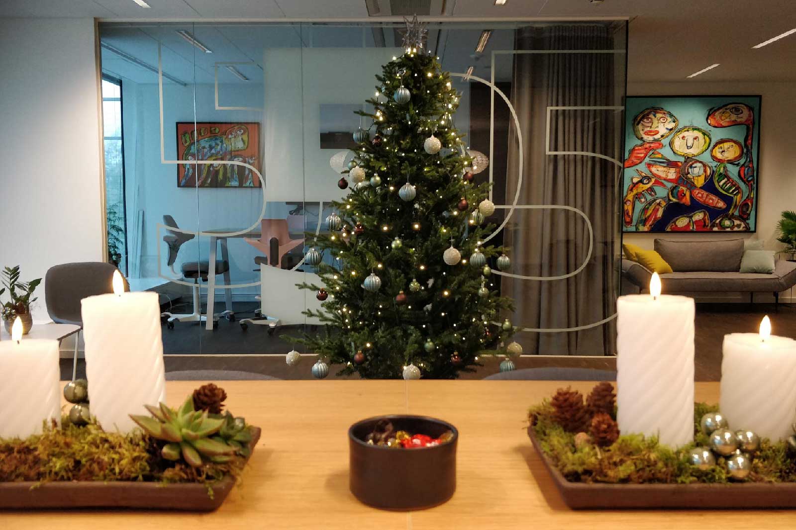 Ein festlich dekorierter Weihnachtsbaum steht im Büro, umgeben von Kerzen und Pflanzen auf einem Holztisch.