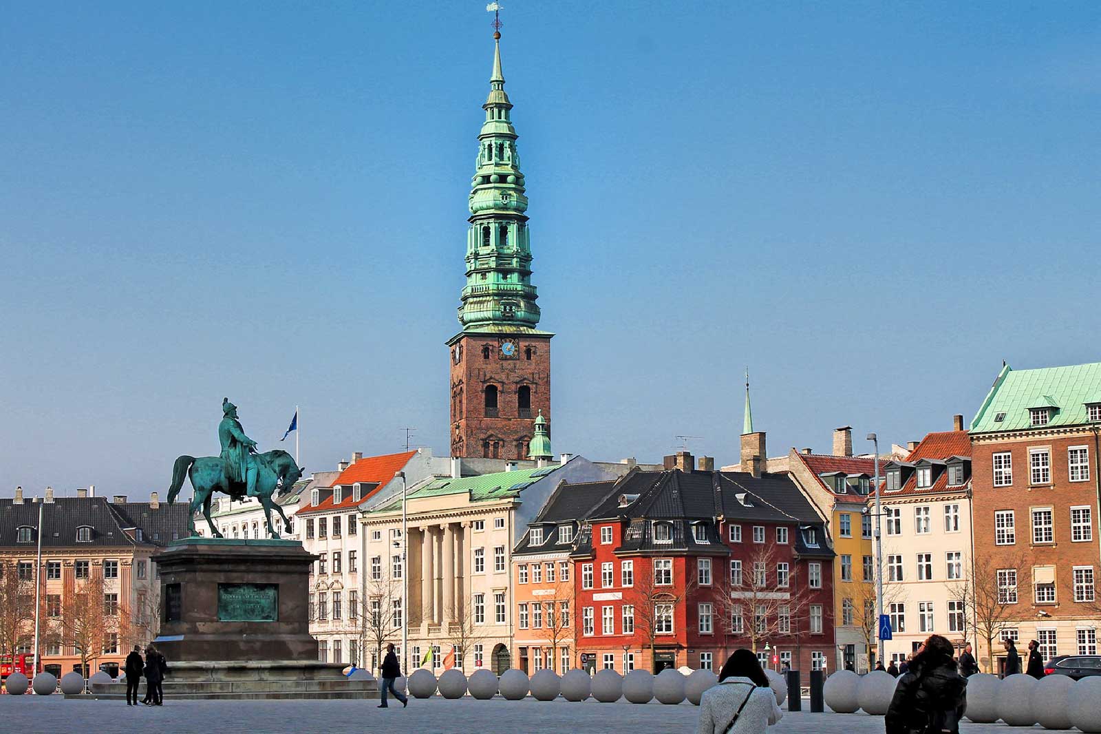 Ein Platz mit einem Reiterdenkmal und bunten Gebäuden. Im Hintergrund eine hohe, grüne Turmspitze unter blauem Himmel.
