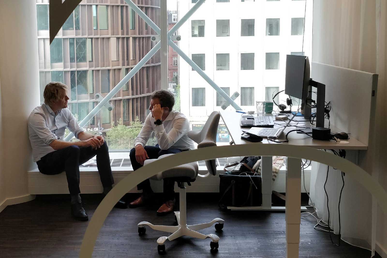 Zwei Männer sitzen in einem modernen Büro mit großen Fenstern und diskutieren. Ein Schreibtisch mit Computer und Büromaterial ist sichtbar.