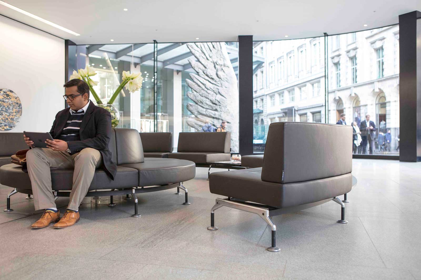 Ein Mann sitzt in einer modernen Lobby auf einem runden Sofa und schaut auf ein Tablet. Im Hintergrund sind große Fenster sichtbar.