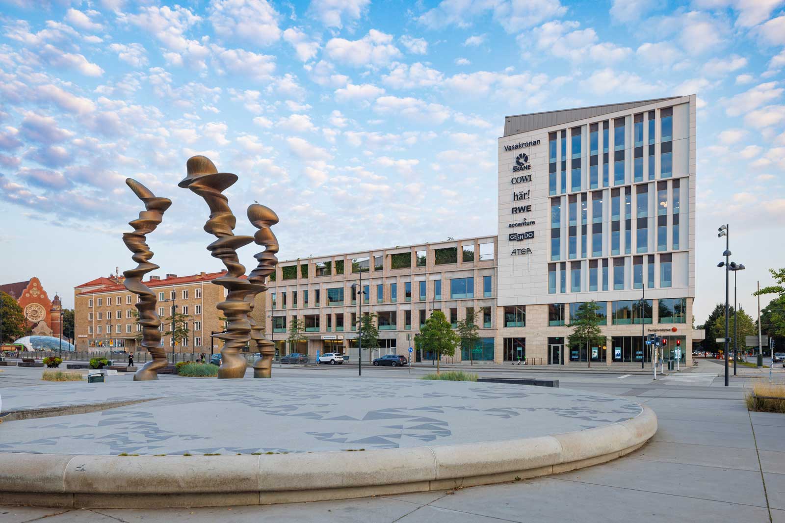Eine moderne Stadtansicht mit einer künstlerischen Skulptur im Vordergrund und modernen Gebäuden im Hintergrund.