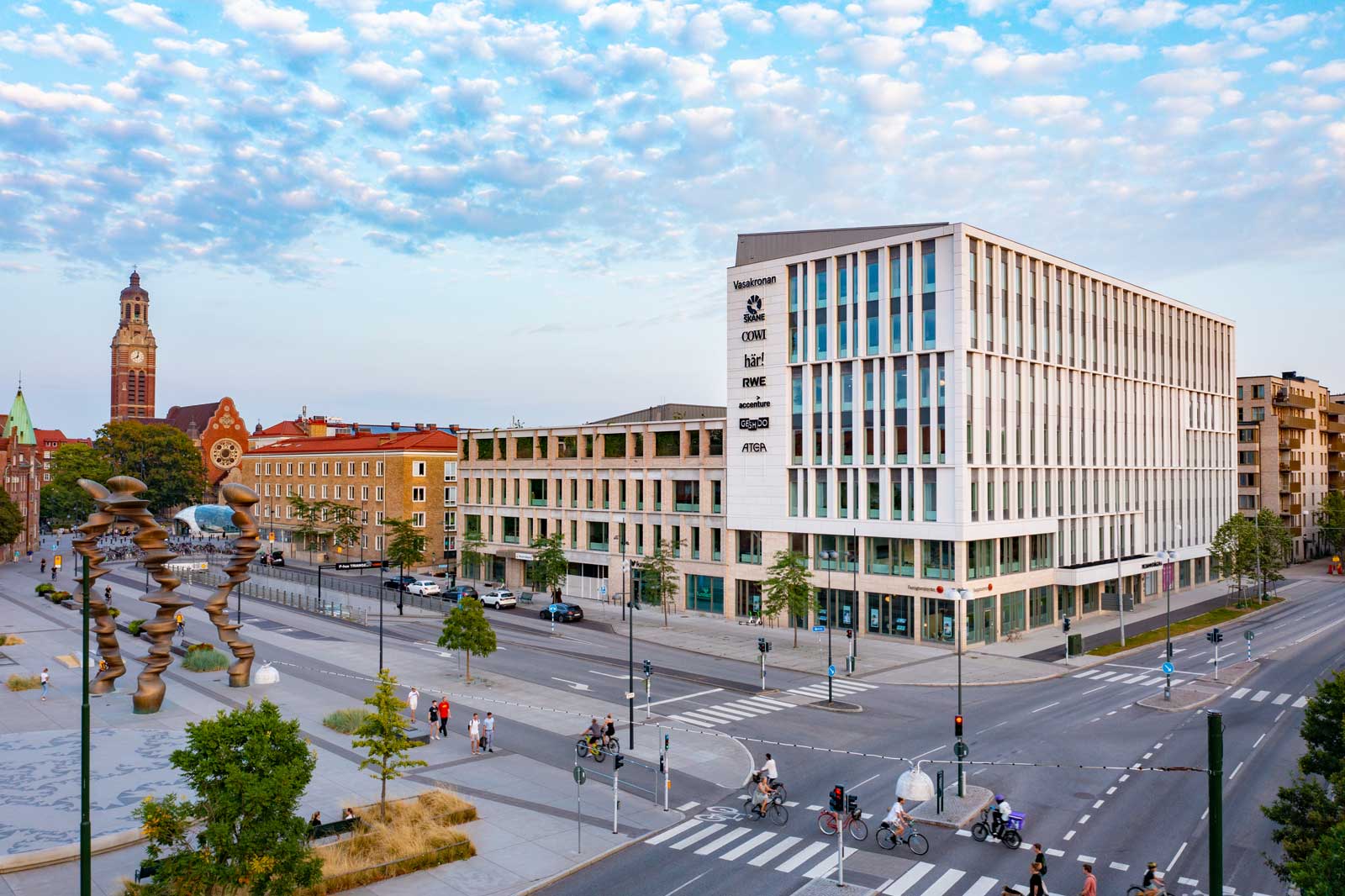 Eine moderne Stadtansicht mit einem neuen Gebäude und einer Uhrturm im Hintergrund. Menschen und Fahrräder sind auf der Straße.