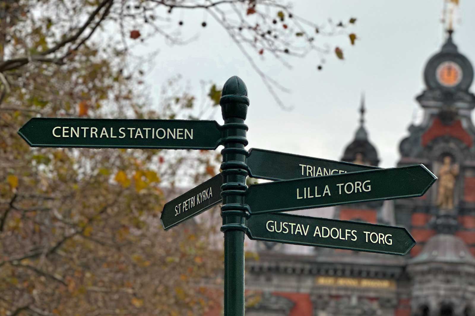 Ein Wegweiser mit Schildern zu verschiedenen Orten: Centralstationen, Triangeln, Lilla Torg und Gustav Adolfs Torg. Im Hintergrund ein historisches Gebäude.