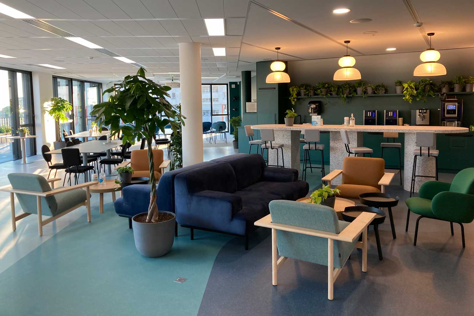 Moderne Lounge mit Pflanzen, Sitzgelegenheiten und einer Bar. Helle Farben und viel Tageslicht schaffen eine einladende Atmosphäre.