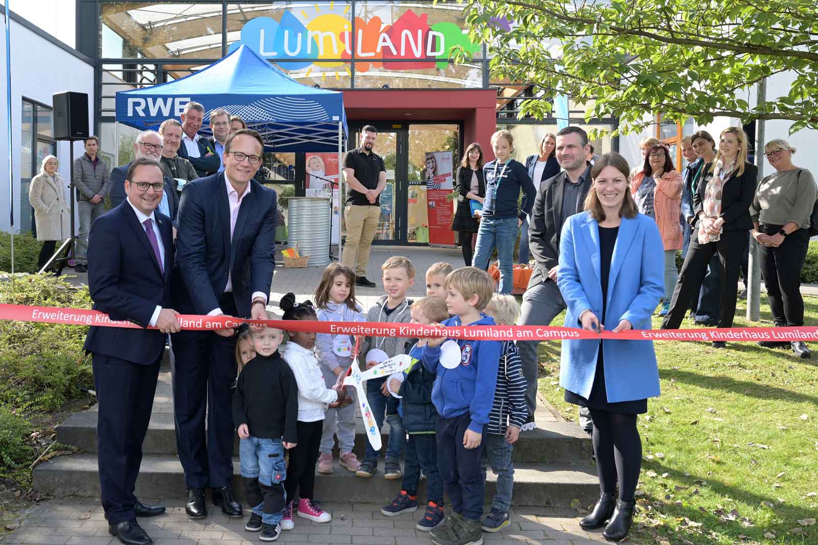 Eine Eröffnungsfeier für Lumiland, mit Kindern und Erwachsenen, die vor dem Gebäude zusammengekommen sind, um die neue Erweiterung zu feiern.