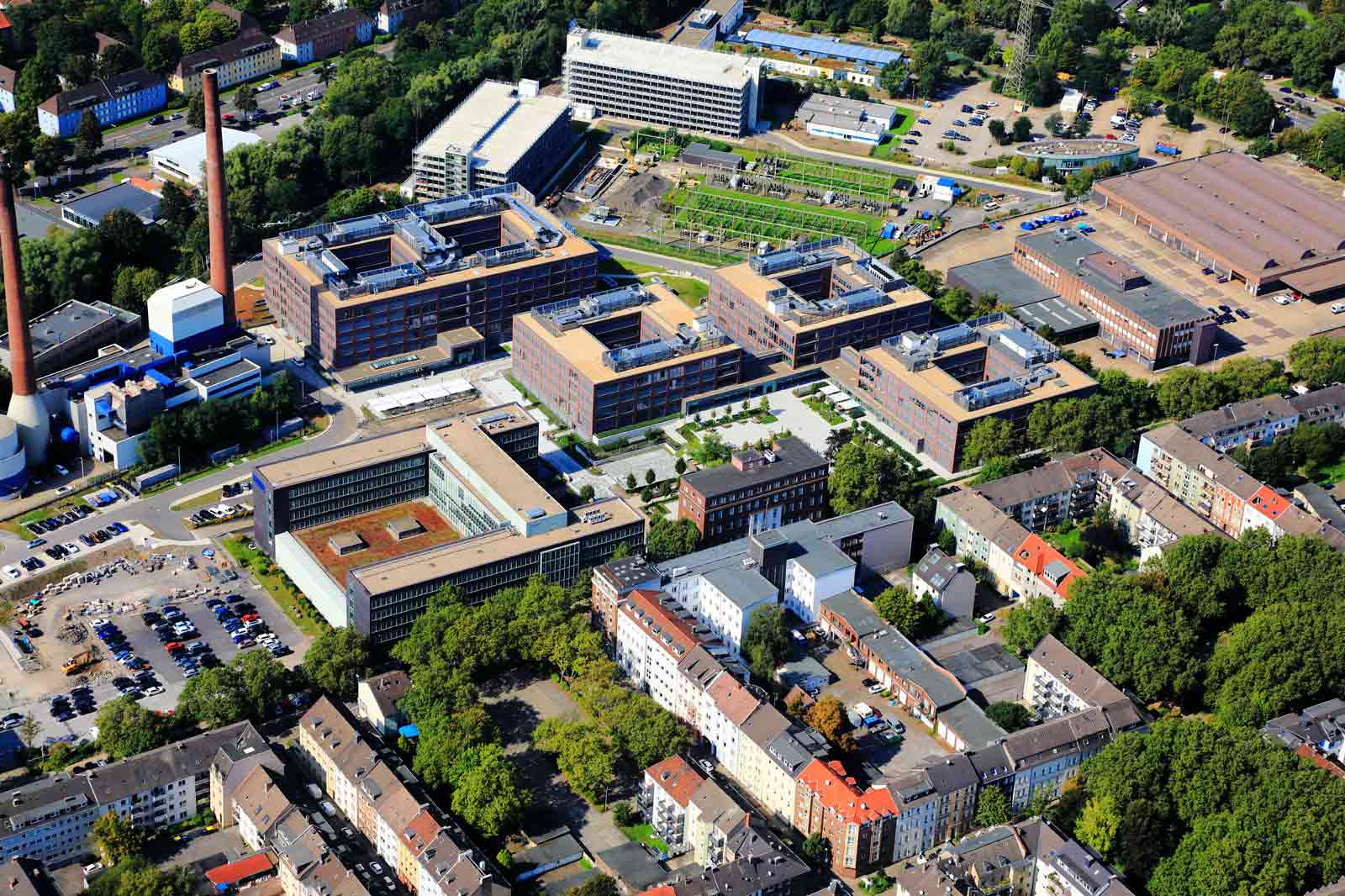 Luftaufnahme eines Komplexes mit modernen Gebäuden, umgeben von Bäumen und Parkplätzen, nahe Wohngebieten.