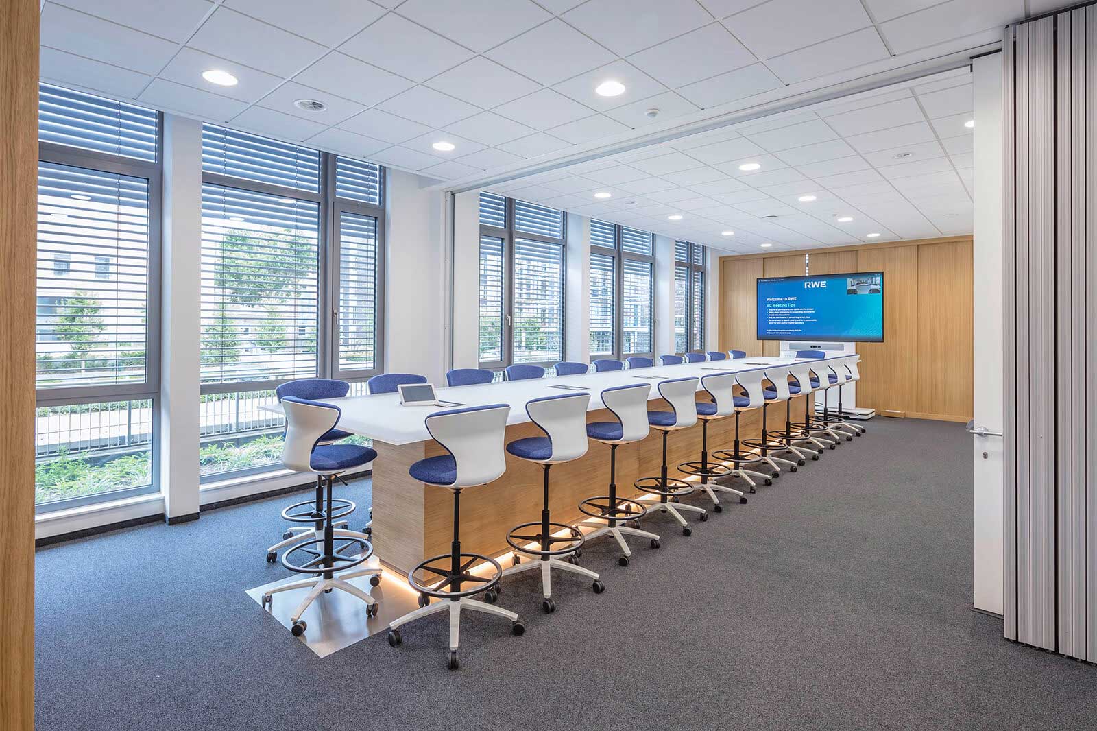 Ein modernes Konferenzzimmer mit einem langen Tisch, blauen Barhockern und großen Fenstern, die viel Tageslicht bieten.