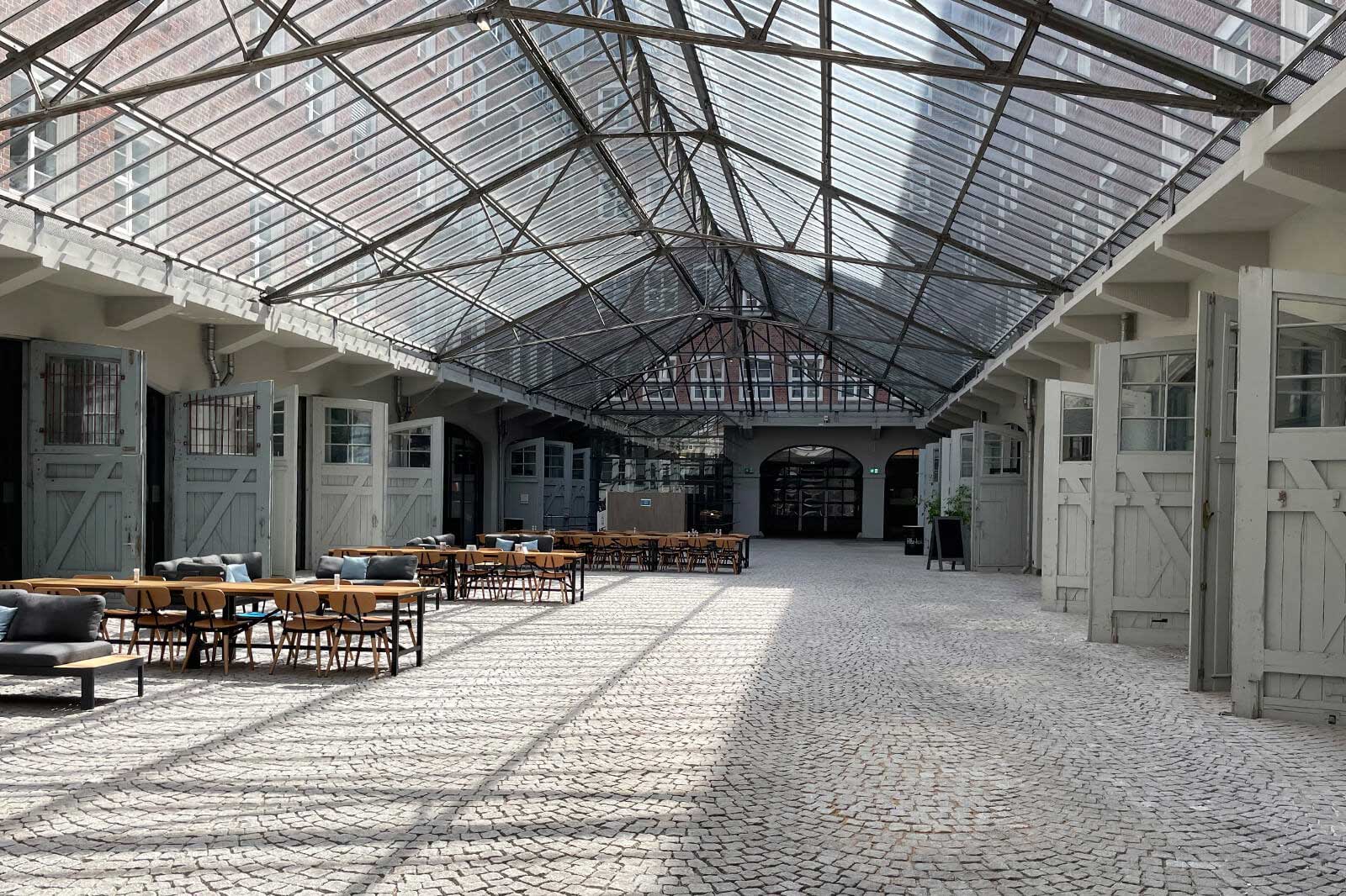 Ein heller, offener Raum mit einer Glasdecke, Tischen und Stühlen. Der Boden ist mit schattierten Steinplatten ausgelegt.