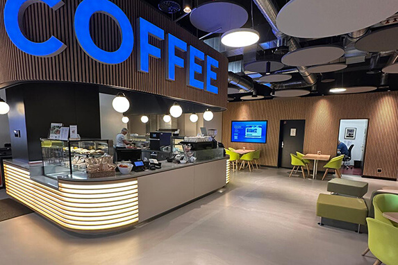 Ein modernes Café mit einer großen blauen 'COFFEE'-Anzeige, Theke mit Snacks und komfortablen Sitzplätzen.
