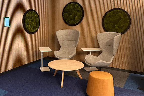 Ein gemütlicher Sitzbereich mit zwei grauen Sesseln, einem runden Tisch und einem orangefarbenen Hocker an einer holzvertäfelten Wand.