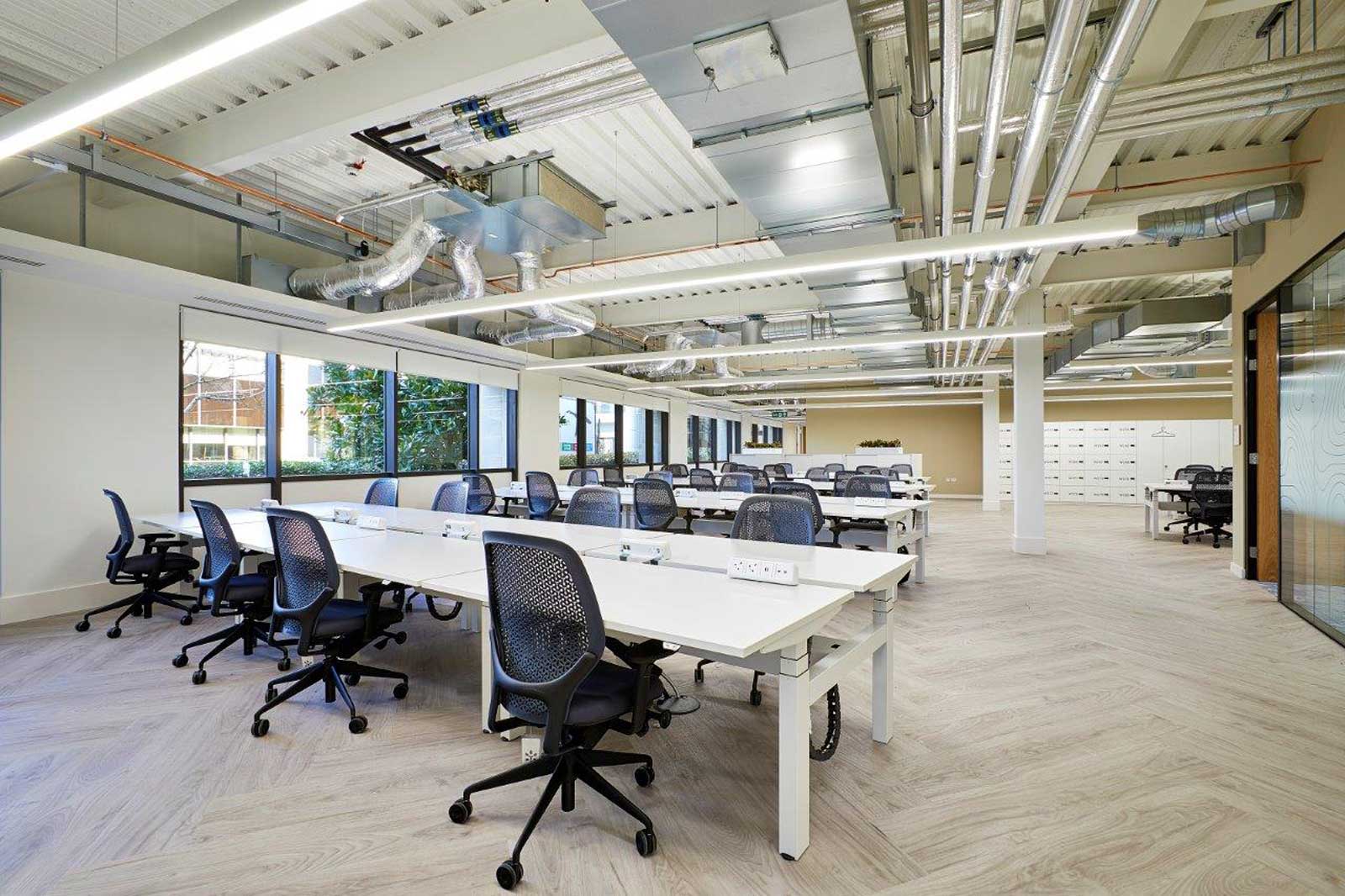 Ein modern gestaltetes Büro mit hellen Tischen, ergonomischen Stühlen und großen Fenstern, die auf einen Garten blicken.