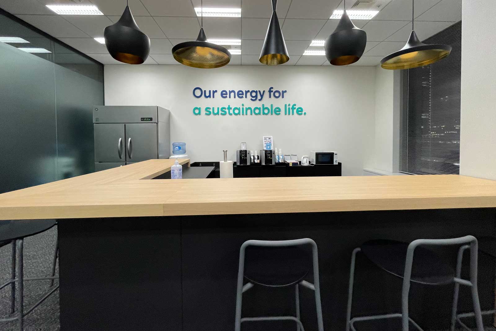 Eine moderne Büroküche mit einer Holzoberfläche, schwarzen Stühlen und einem Wand-Slogan über nachhaltige Energie.