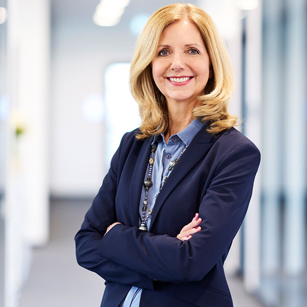 Eine Frau in einem blauen Anzug steht selbstbewusst mit verschränkten Armen in einem modernen Büro.