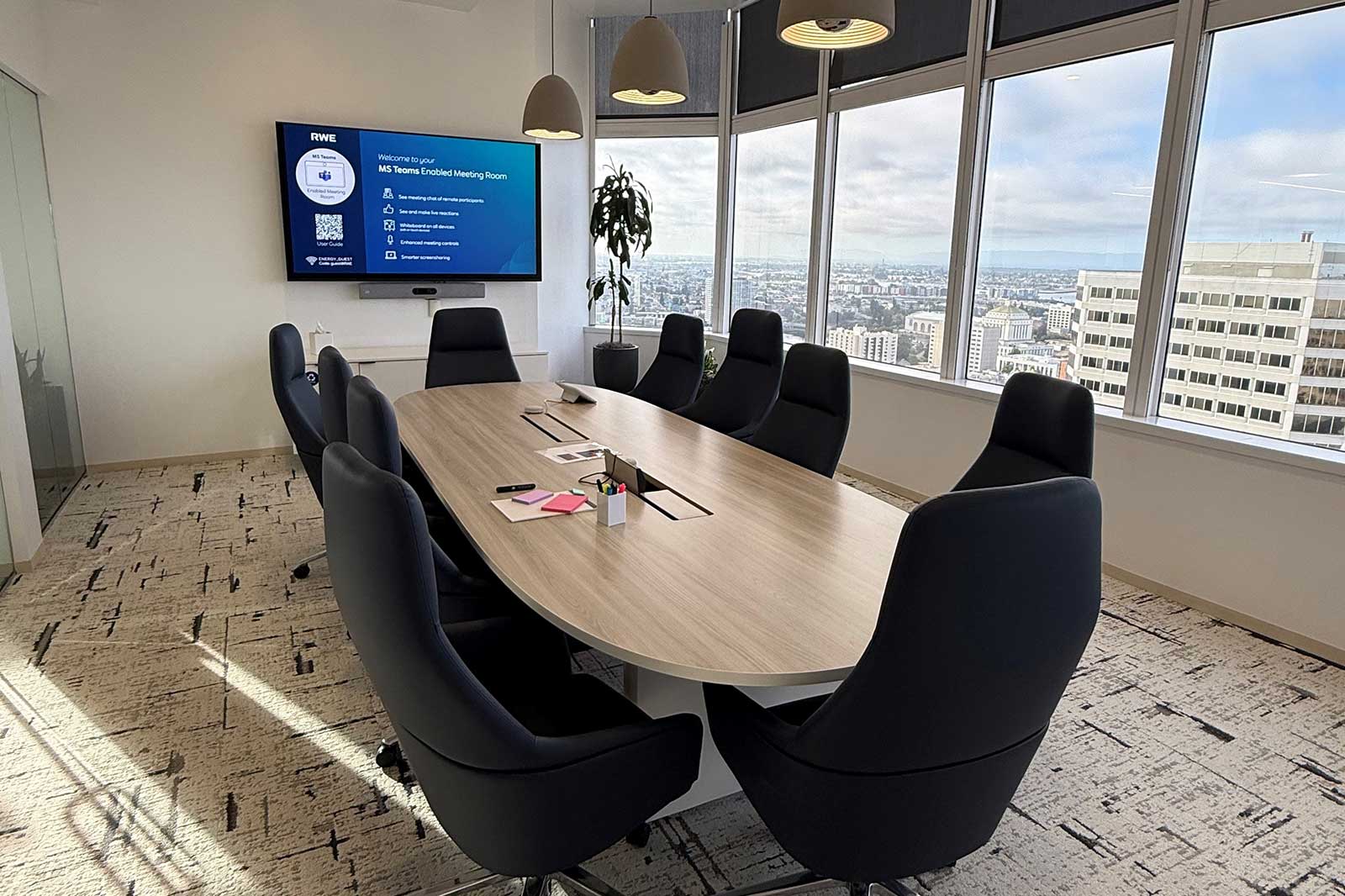 A modern meeting room featuring a large oval table with black chairs, a digital screen, and large windows displaying a city view.