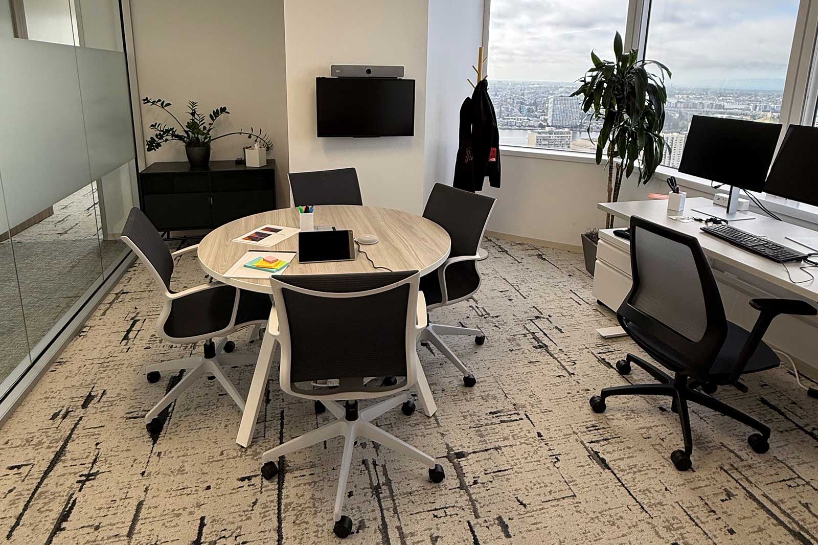 A modern conference room featuring a round table with six chairs, a plant, and large windows overlooking the city skyline.