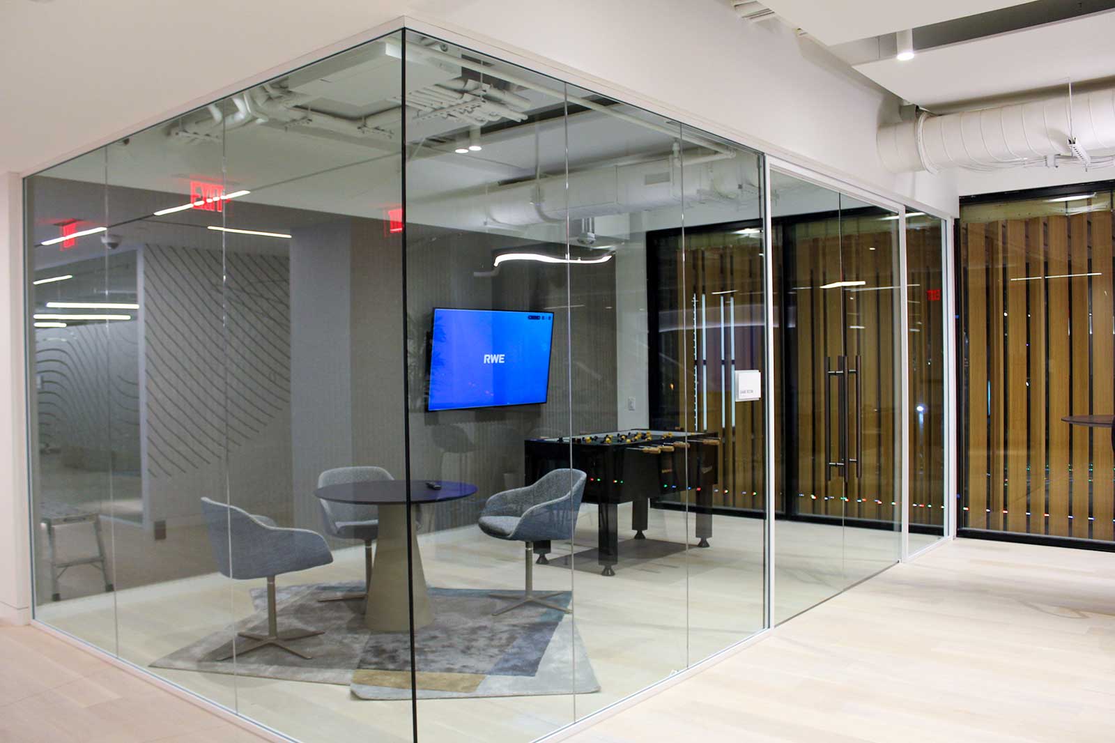 A modern glass meeting room featuring two grey chairs, a round table, and a screen displaying 'RWE'. A foosball table is visible.