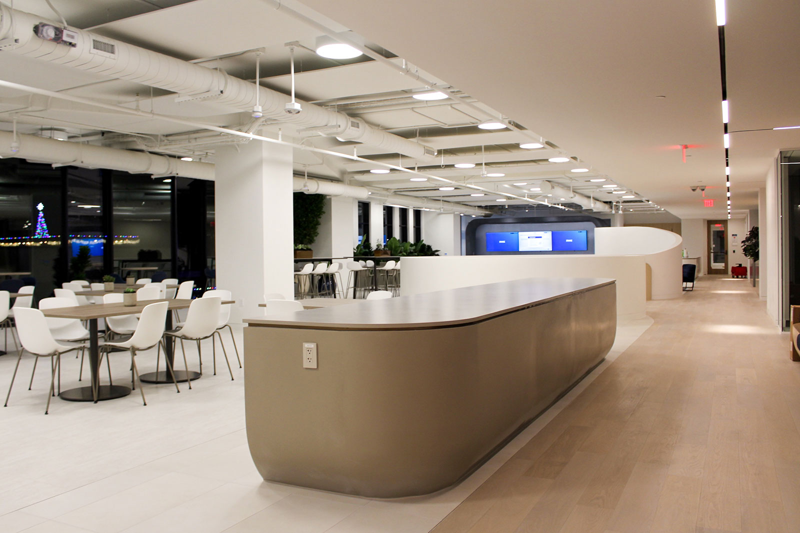 A modern office interior featuring a sleek counter, dining tables with chairs, and a large display screen.